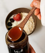 Wooden spoon with honey over a jar of honey, with a blurred background of a bowl with fruits and grains.
