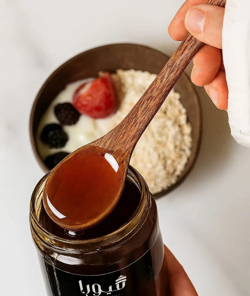 Wooden spoon with honey over a jar of honey, with a blurred background of a bowl with fruits and grains.